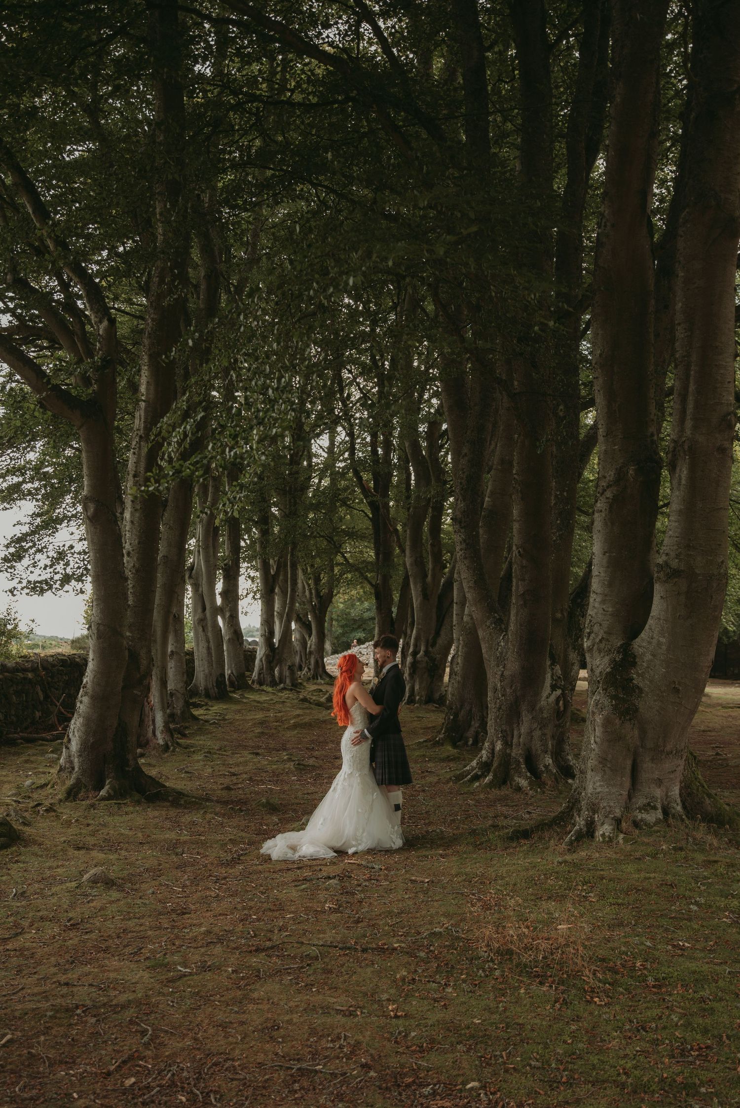 Bride in white gown and groom in tartan kilt embrace in a dramatic tree-lined avenue.
