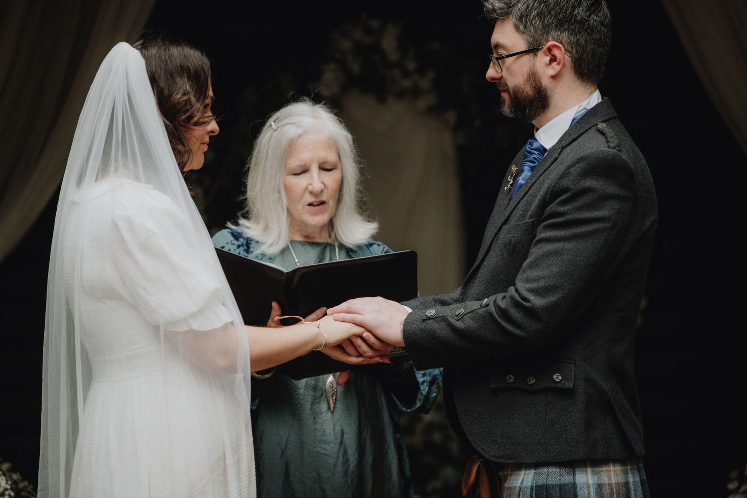 Officiant in teal dress joins bride and kilted groom in hand-holding vow exchange at woodland wedding.
