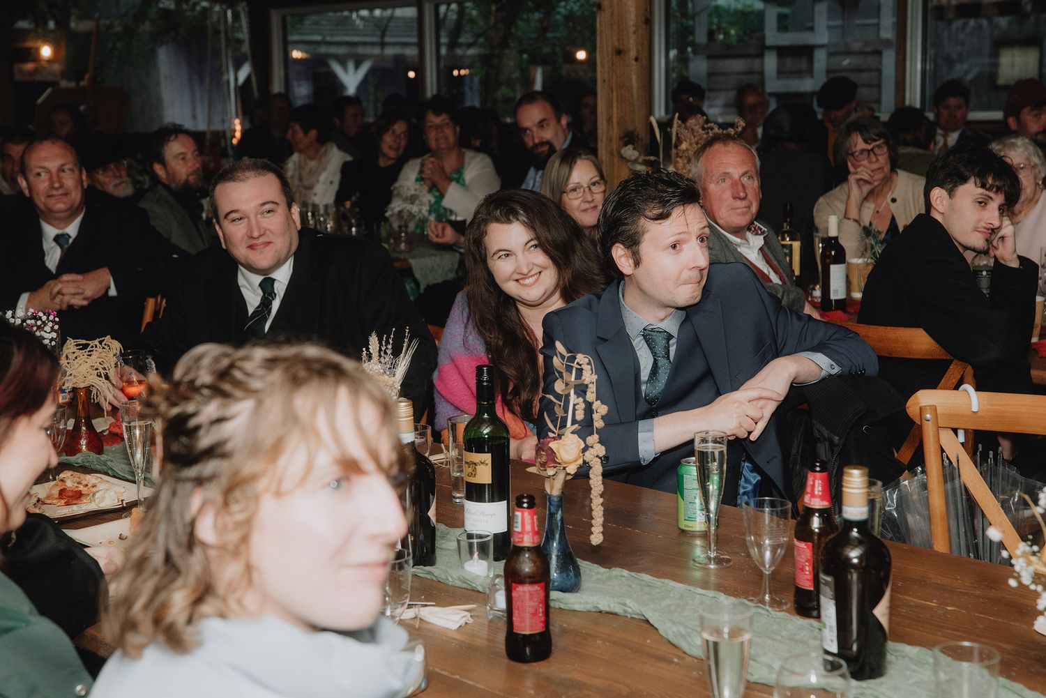 Guests seated at long tables with wine bottles enjoying a wedding reception dinner in a warmly lit restaurant.