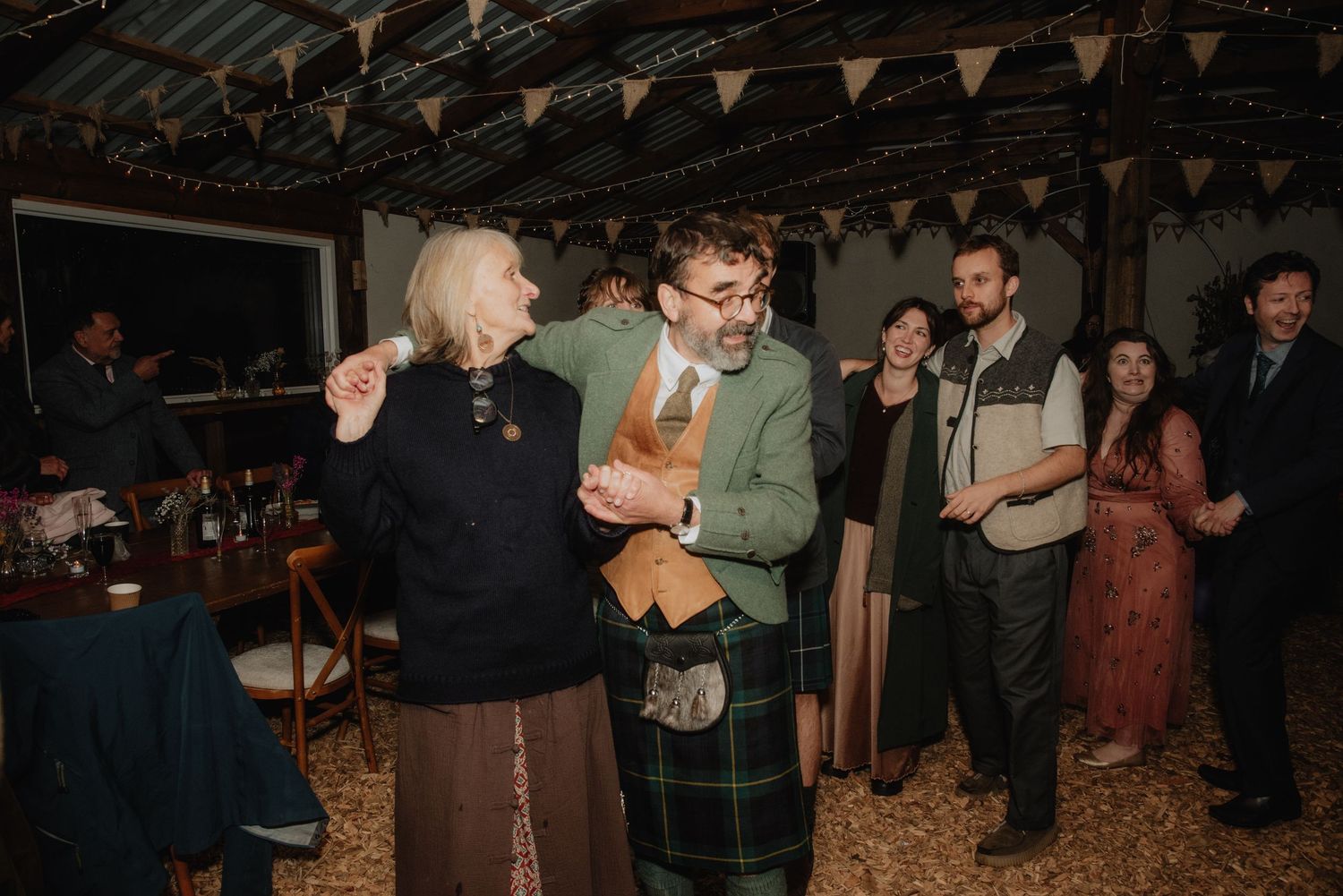 Man in tartan kilt dancing with elderly woman at a Scottish wedding celebration with guests watching.