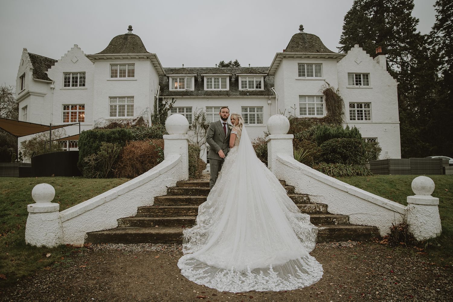 Bride and groom pose on white stone steps of elegant manor house wedding venue, bride's lace train cascading down stairs.