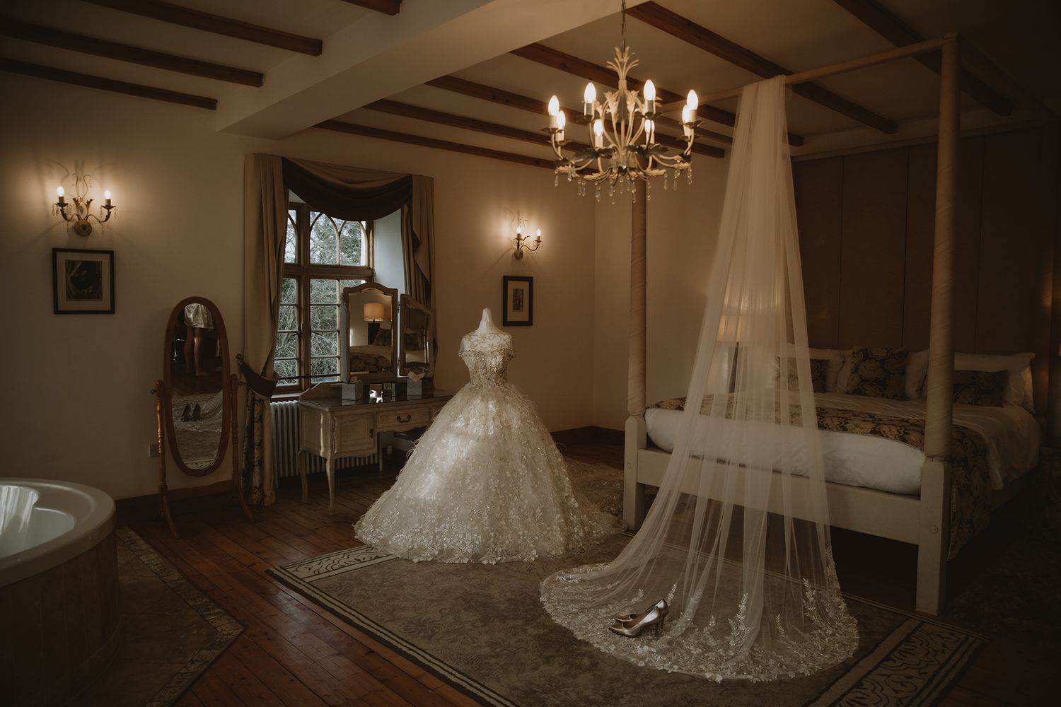 Elegant wedding dress on mannequin in luxurious bedroom with chandelier, four-poster bed, and long veil trailing on floor.