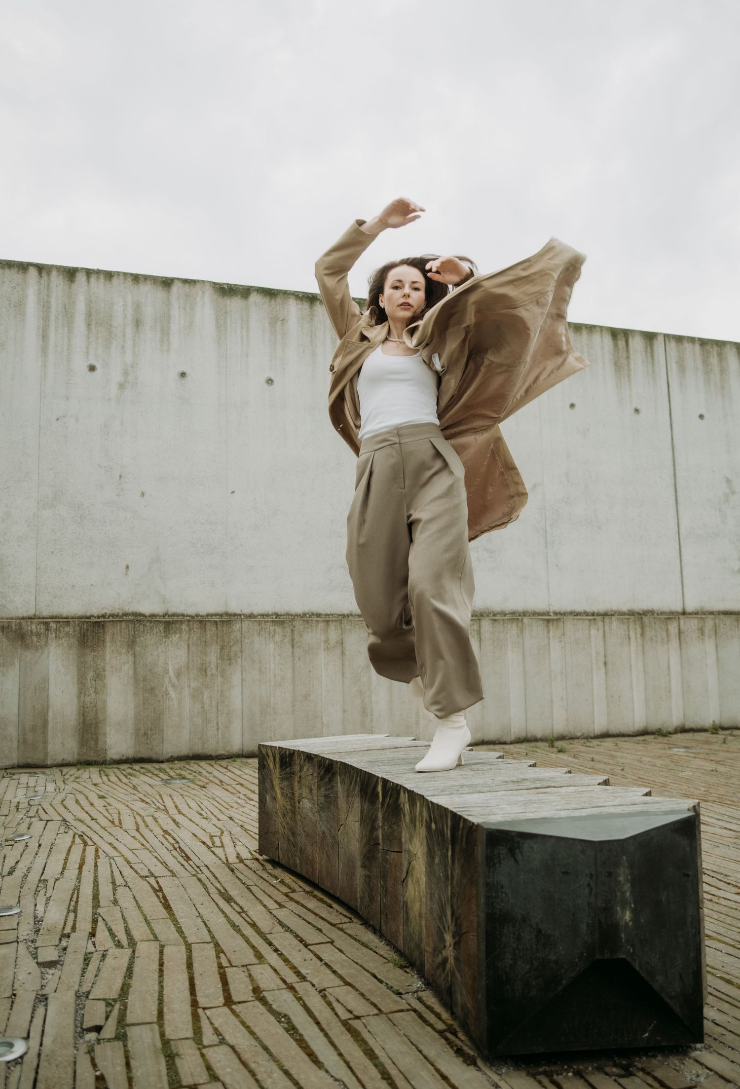 Woman in beige wide-leg pants and brown coat jumps energetically on a concrete bench outdoors.