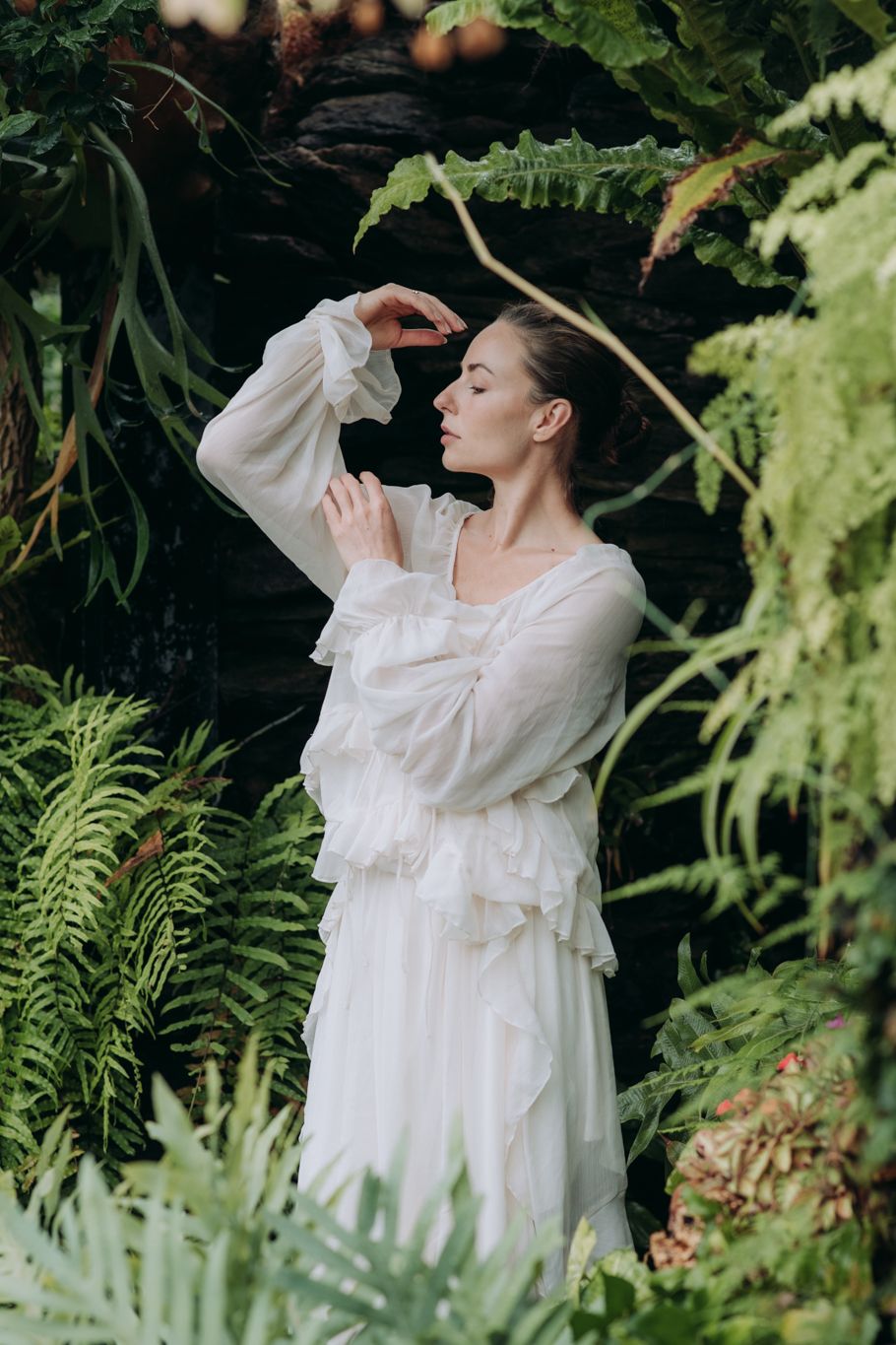 Woman in white ruffled dress poses gracefully among lush green ferns in a forest setting.