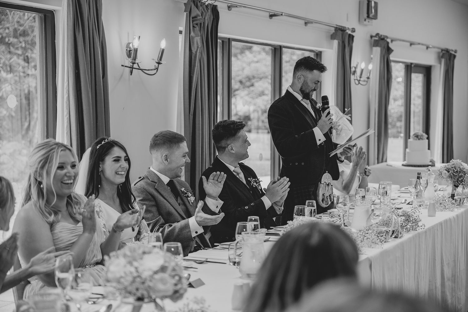 Black and white wedding reception photo of guests laughing during a groom's speech at an elegant venue.