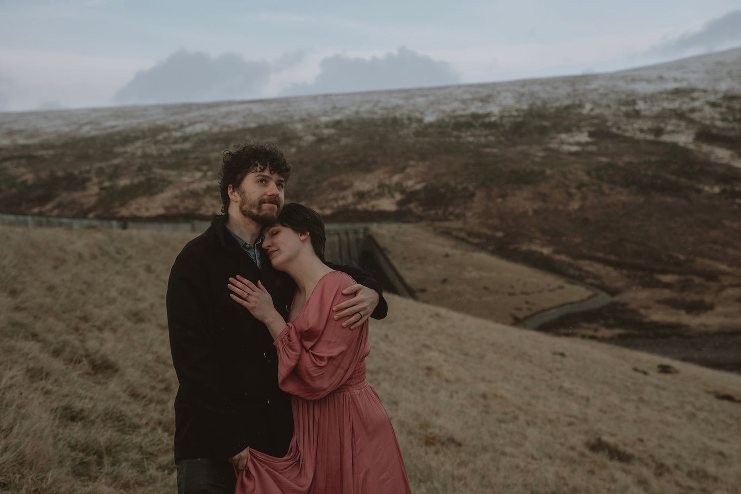 A couple embraces romantically on a misty highland landscape with dramatic mountains in the background.