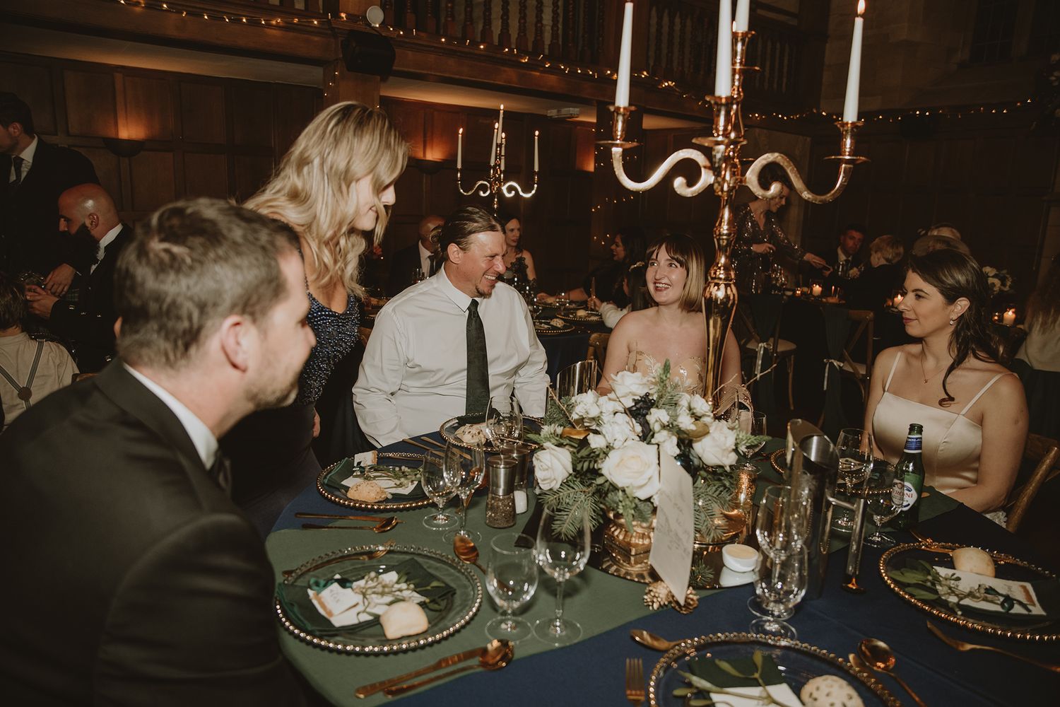 Elegant wedding reception with guests gathered around a navy-draped table adorned with candelabras and white floral centerpieces.