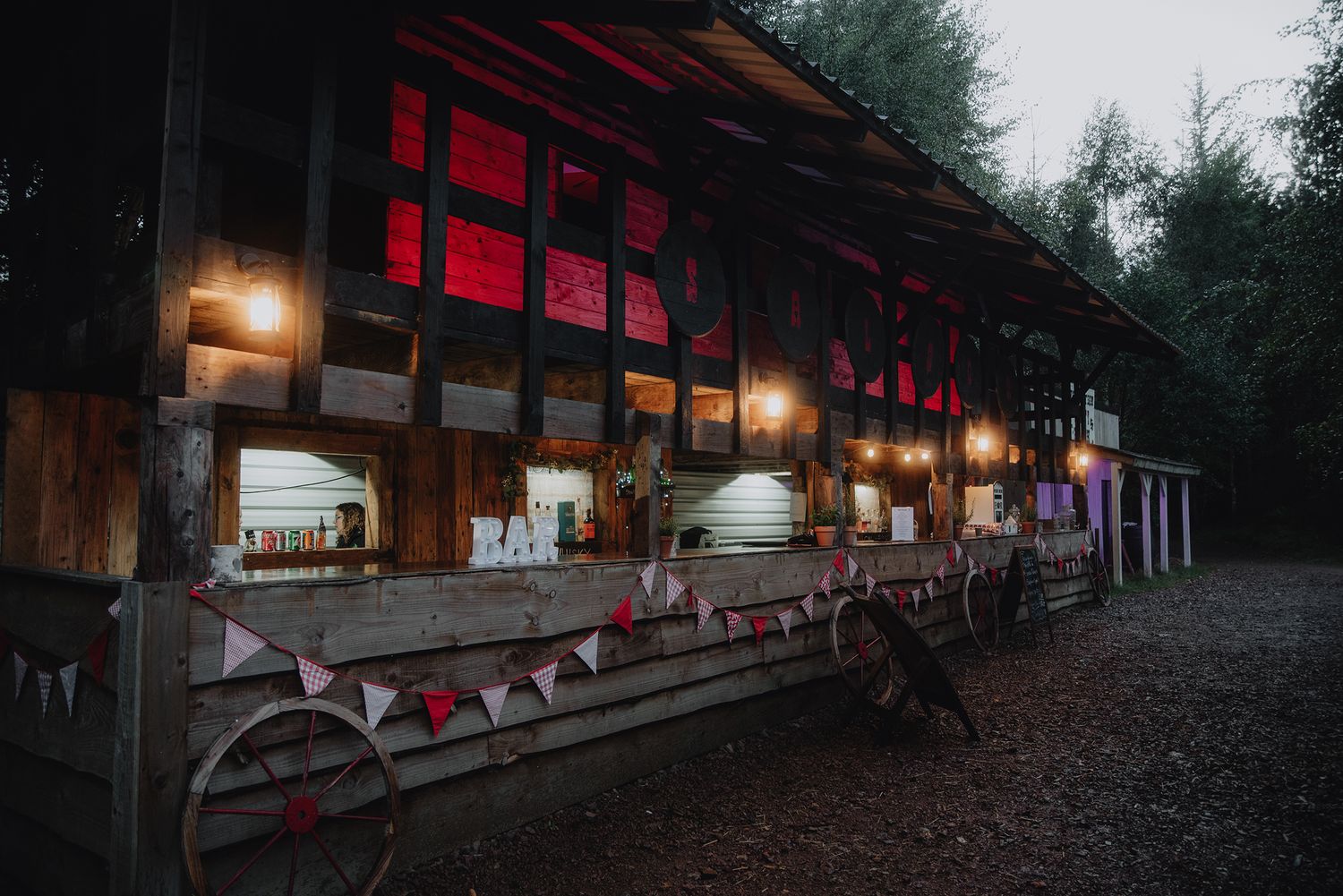 Rustic wooden barn wedding venue exterior lit with warm string lights and bunting at dusk in a forest setting.