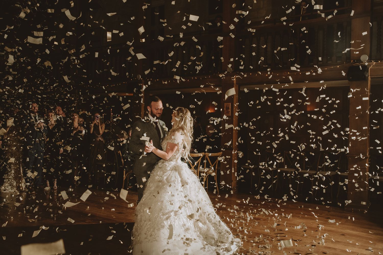 Bride and groom share first dance surrounded by falling gold confetti in a romantic dimly lit wedding venue.