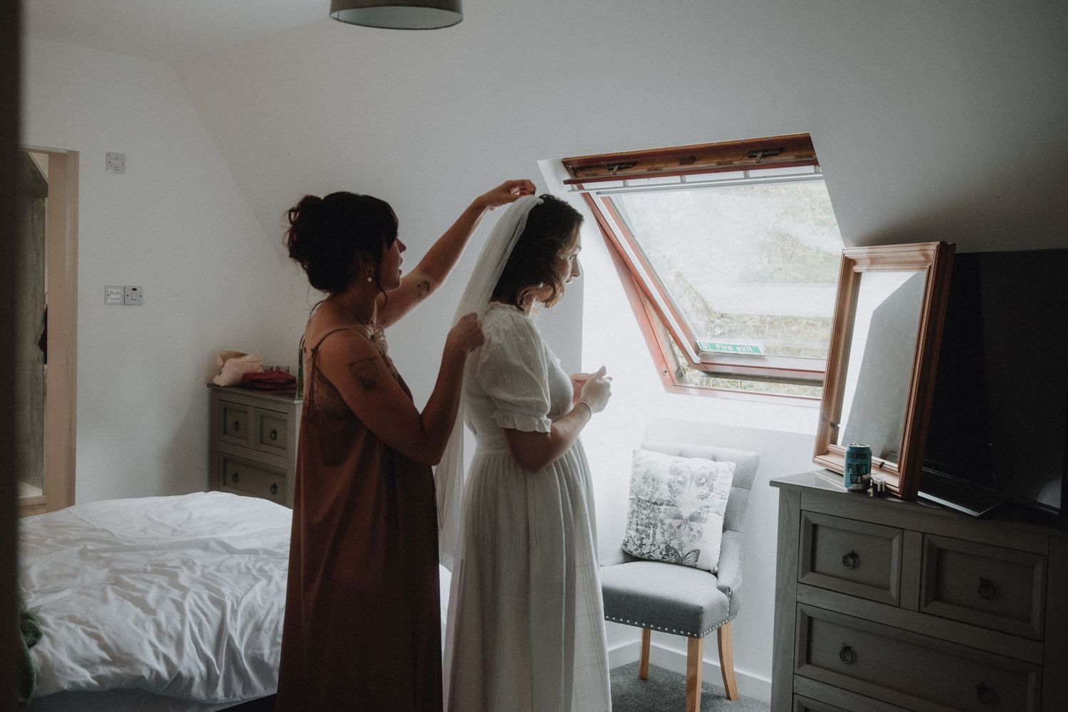 Bridesmaid lifting bridal veil over bride in white dress in bright attic bedroom before wedding ceremony.