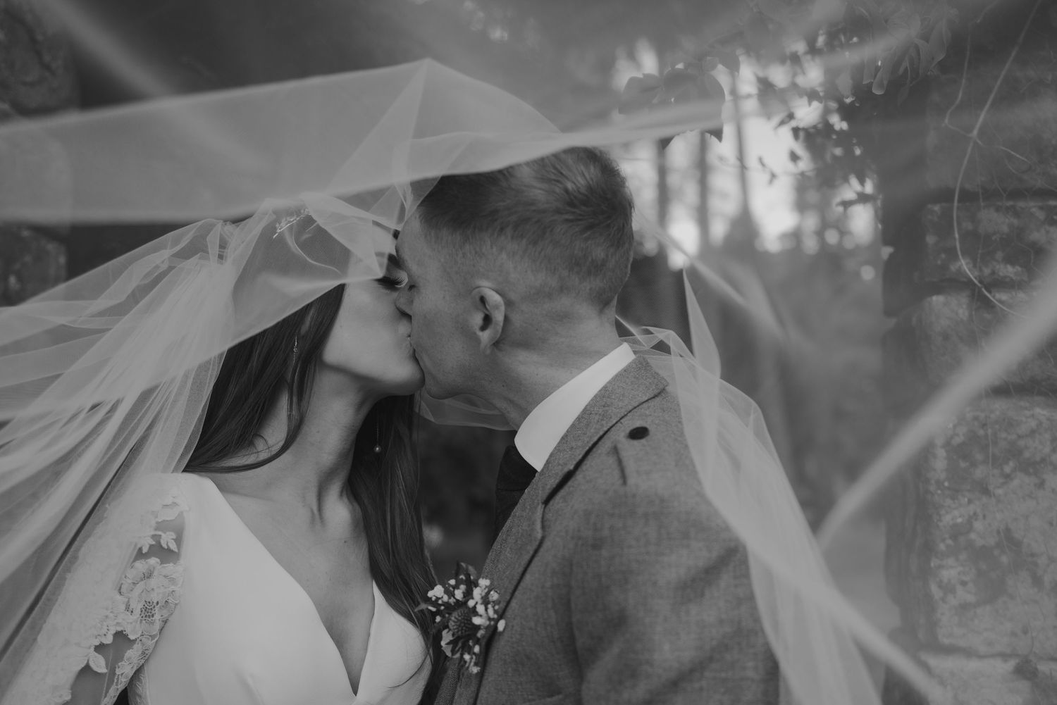 Black and white photo of bride and groom kissing under a wedding veil in a romantic outdoor setting.