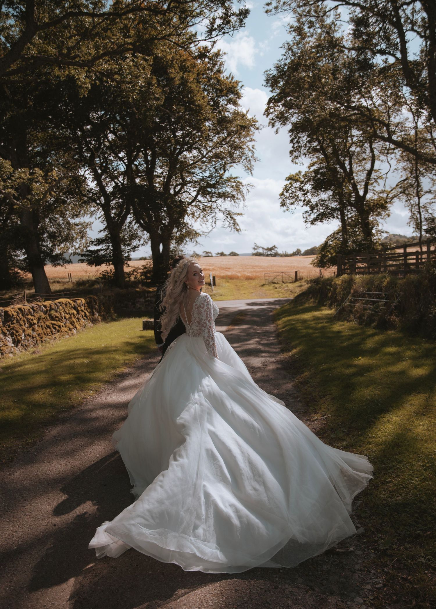 Bride in flowing white ballgown twirling on a tree-lined country lane under a bright blue sky.