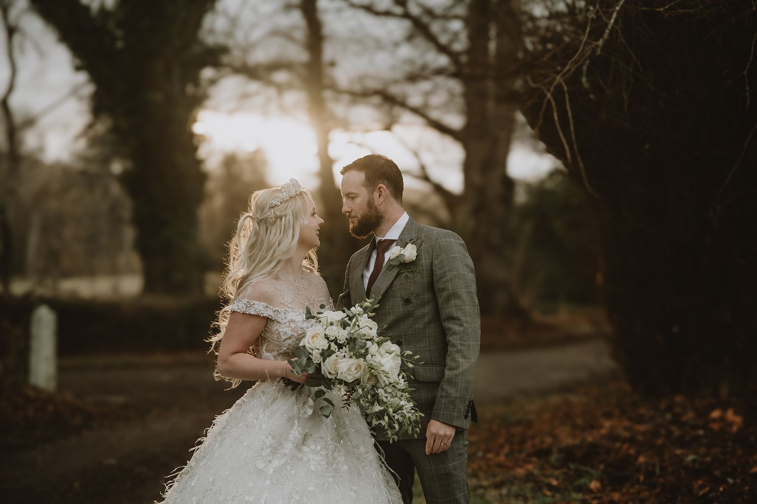 Bride and groom share a romantic moment outdoors, bride holding white floral bouquet, golden sunset light filtering through bare trees.