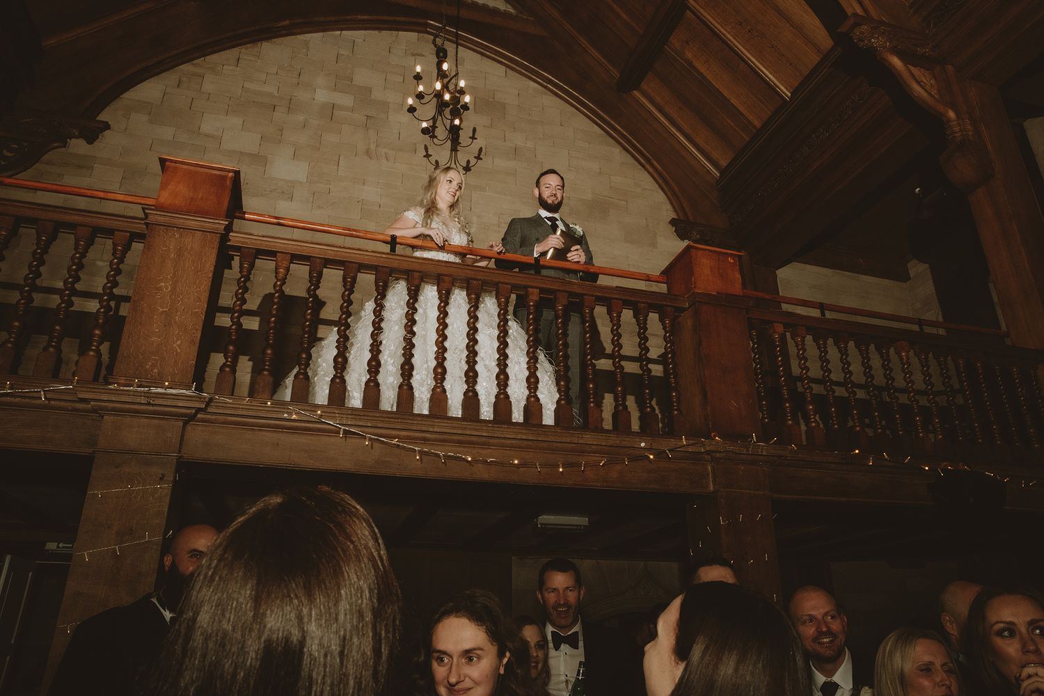 Bride and groom stand on a wooden balcony overlooking wedding guests in an elegant stone venue.