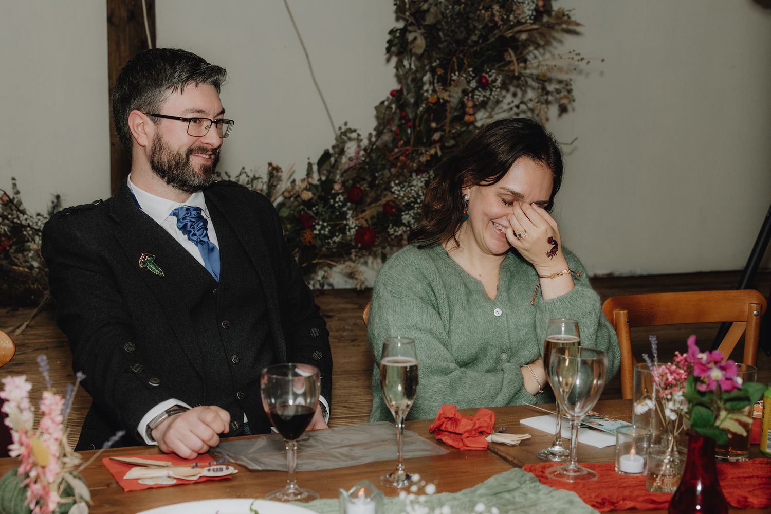 Couple laughing and smiling at wedding reception table with wine glasses and floral decor.