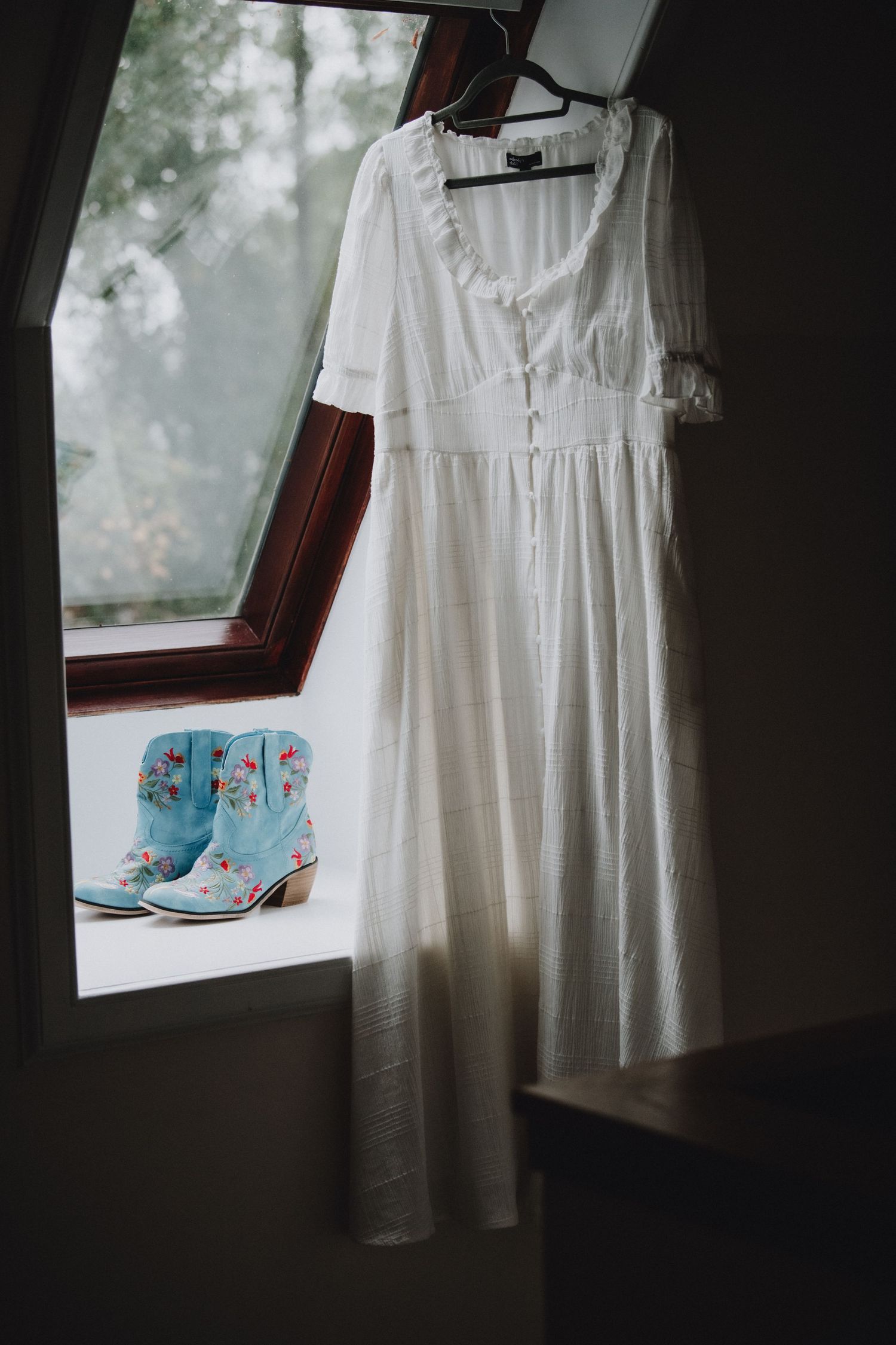 White wedding dress hangs by window beside blue floral embroidered cowboy boots on rustic windowsill.