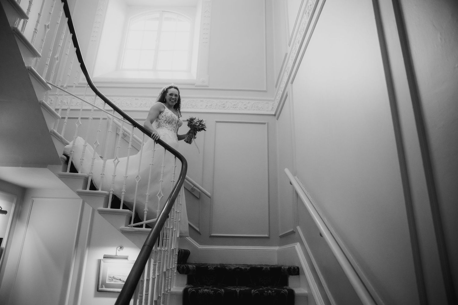 Elegant bride in white wedding dress descends grand staircase holding bouquet in classic black and white photo.