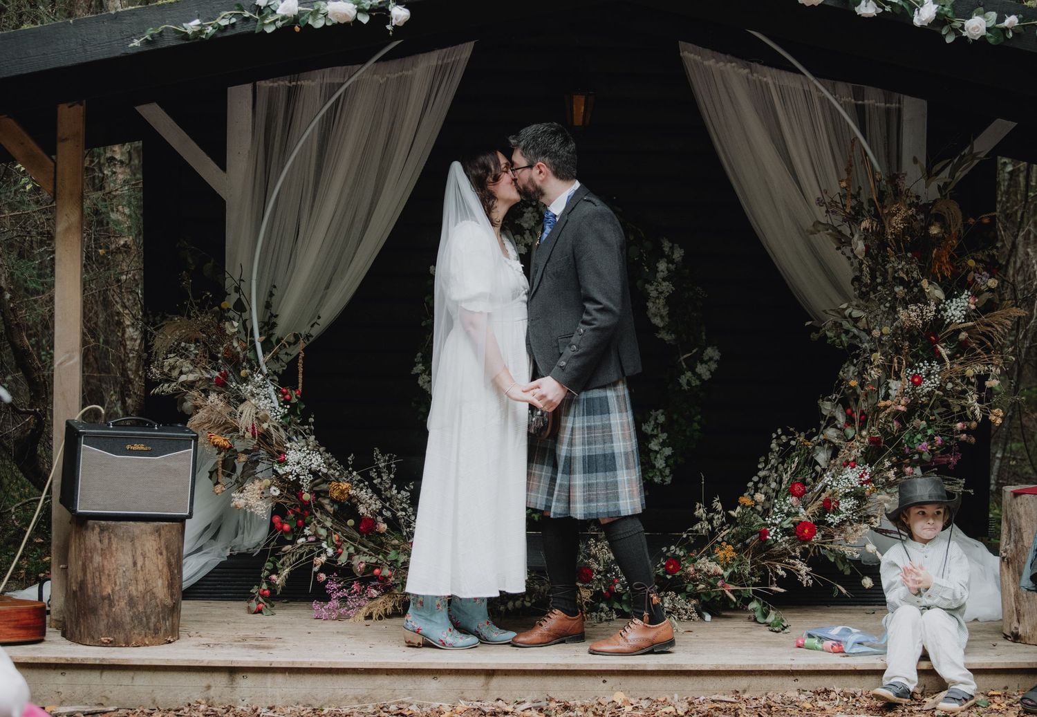 Bride and groom share first kiss at rustic outdoor woodland wedding ceremony.