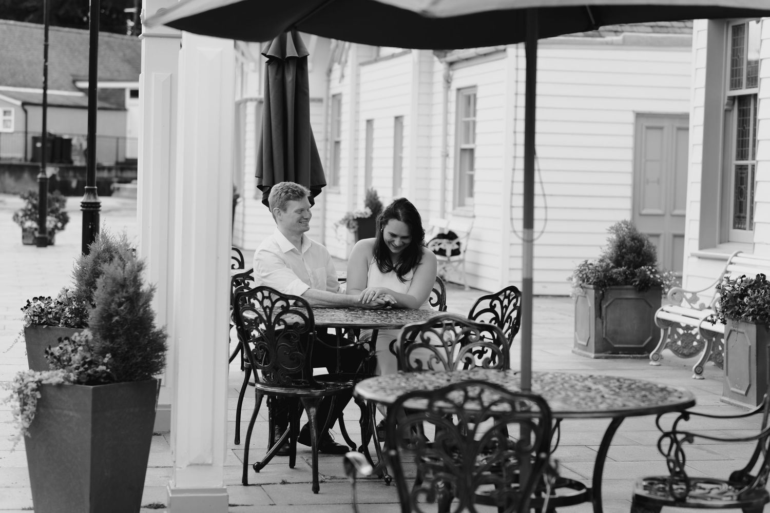 Couple sharing a romantic moment at an outdoor cafe with ornate iron chairs and lush potted plants.