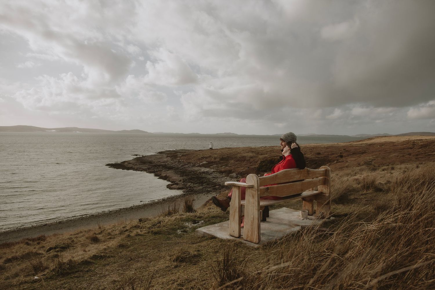 Person sitting alone on wooden bench overlooking stormy Scottish coastal landscape.