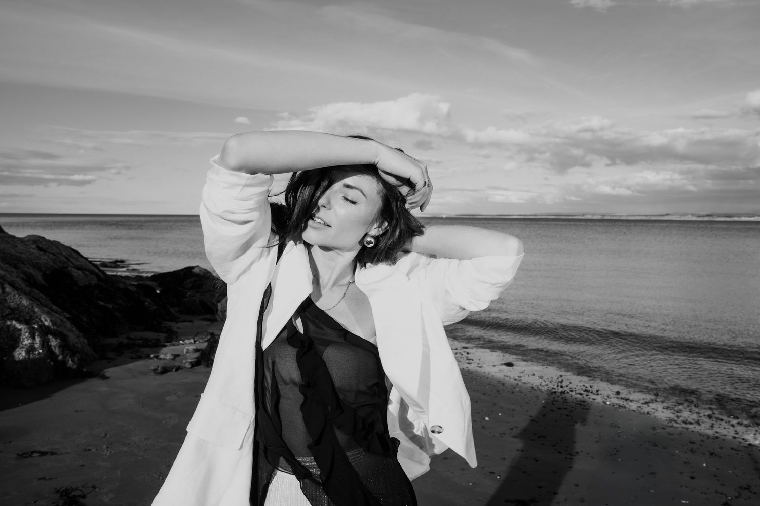Black and white photo of a woman in white jacket posing on a windy beach at sunset.