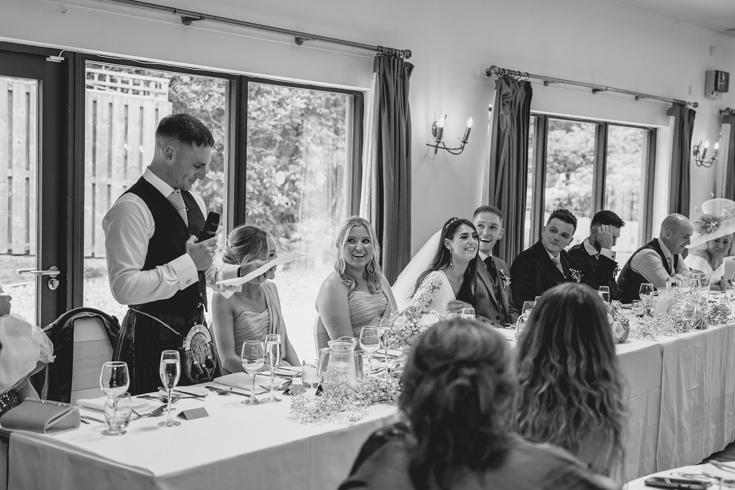 Black and white wedding reception photo showing groom giving speech at head table with smiling bride and guests.