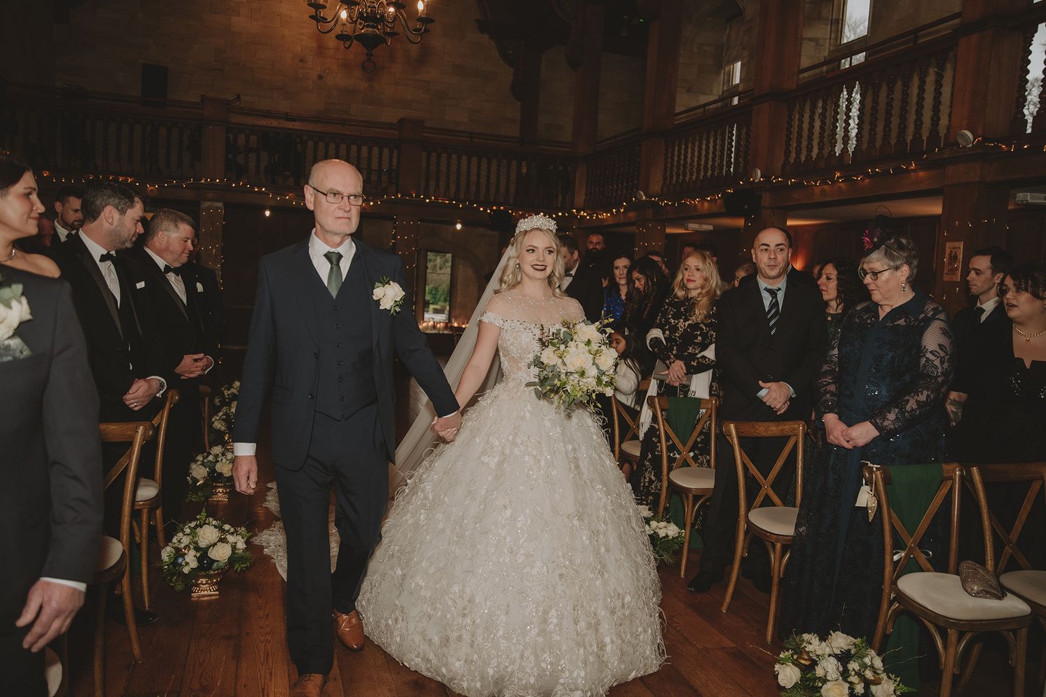 Bride in ball gown walks down aisle with escort in historic venue during elegant wedding ceremony.