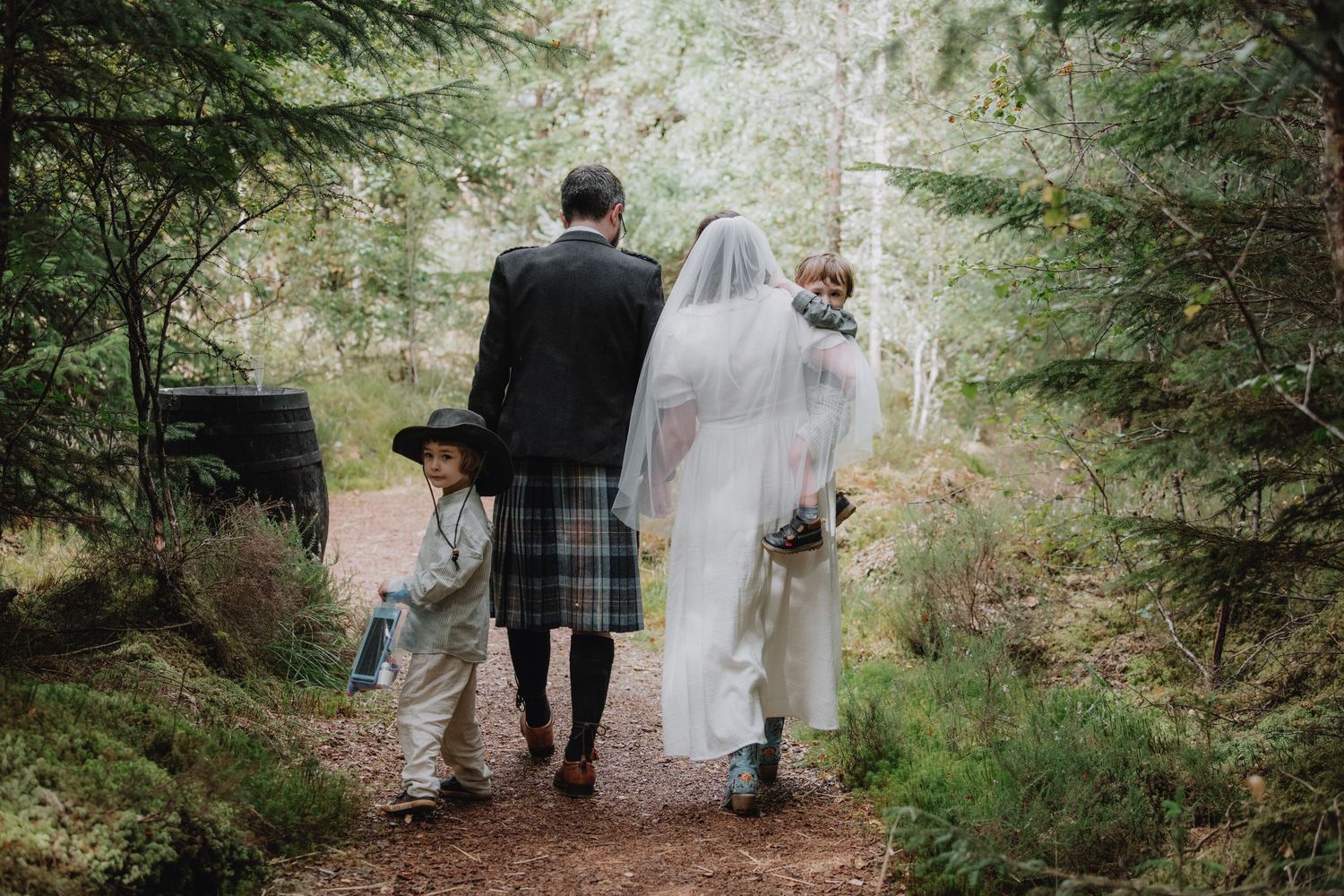 Wedding couple in white dress and tartan kilt stroll forest trail with children in woodland setting.