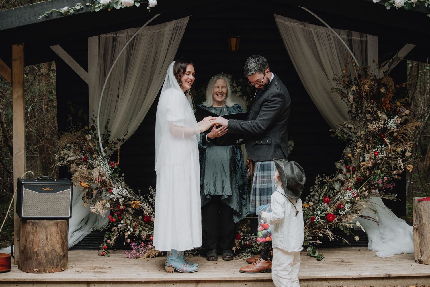 Smiling bride and kilted groom exchange rings before officiant at decorated outdoor woodland wedding ceremony.