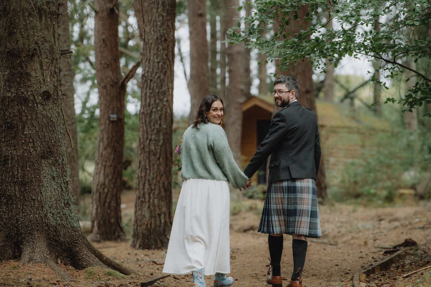 A couple in wedding attire walk hand in hand through a pine forest, groom wearing a Scottish kilt.