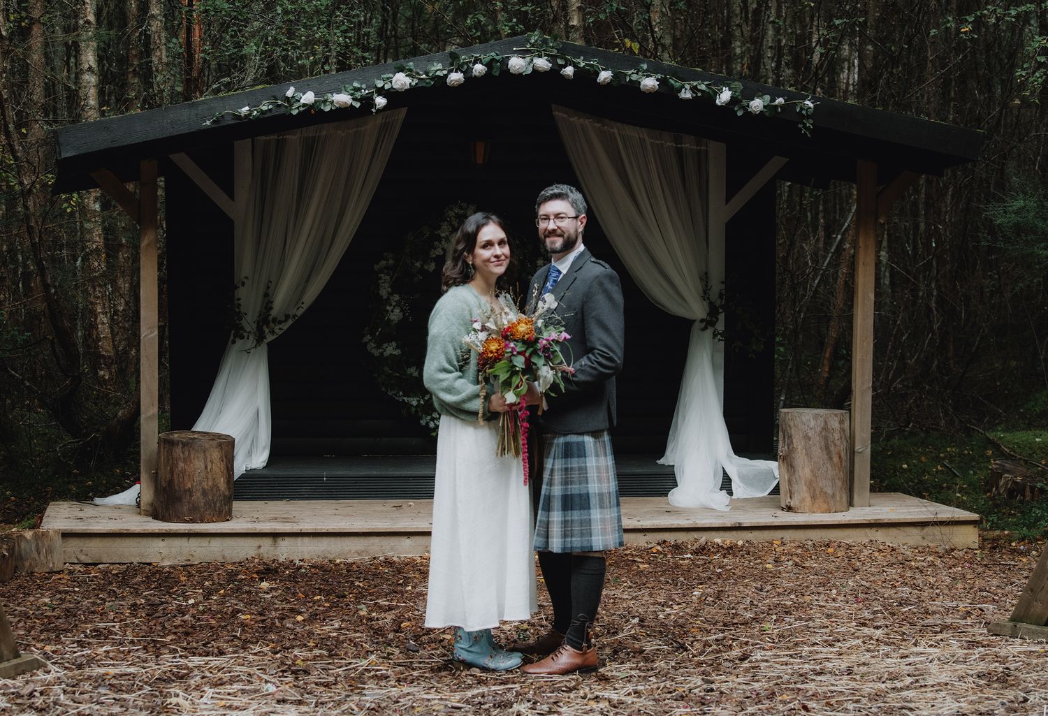 A smiling bride and groom pose before a draped woodland wedding ceremony arch in a forest.