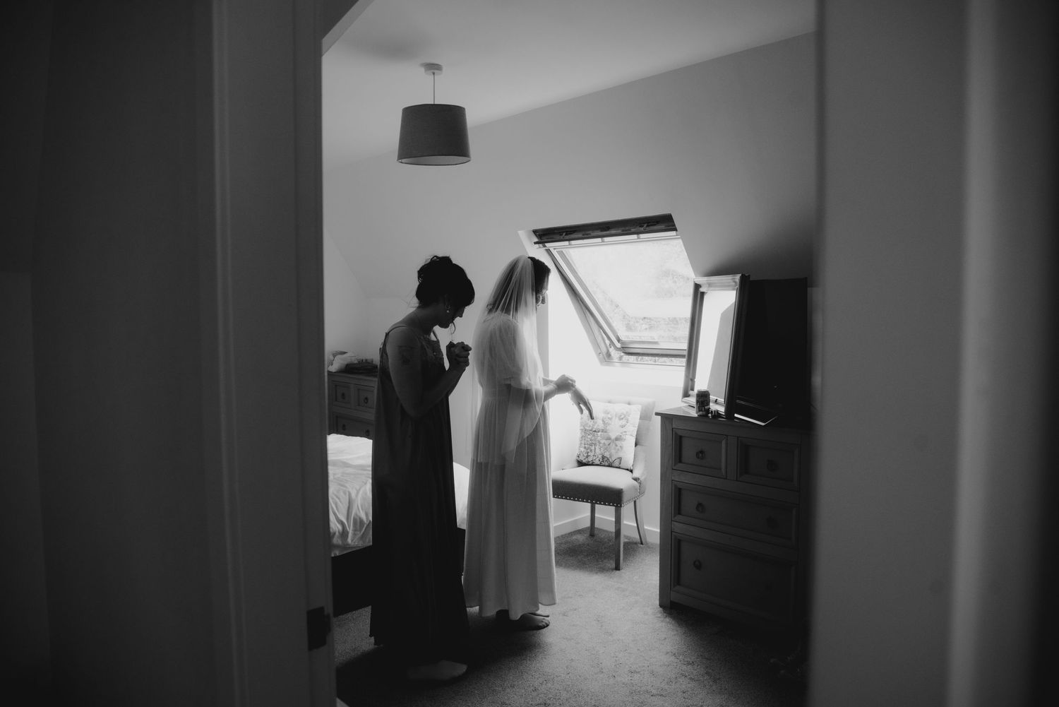 Black and white photo of bride getting veil fitted by bridesmaid in a cozy attic bedroom before wedding.