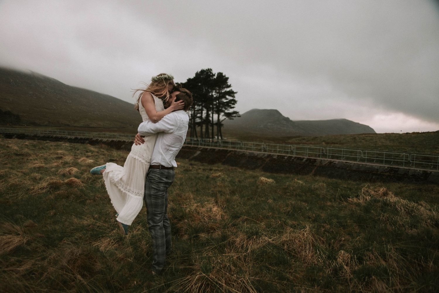 Man lifting and kissing woman in dramatic Scottish Highland landscape under stormy skies.