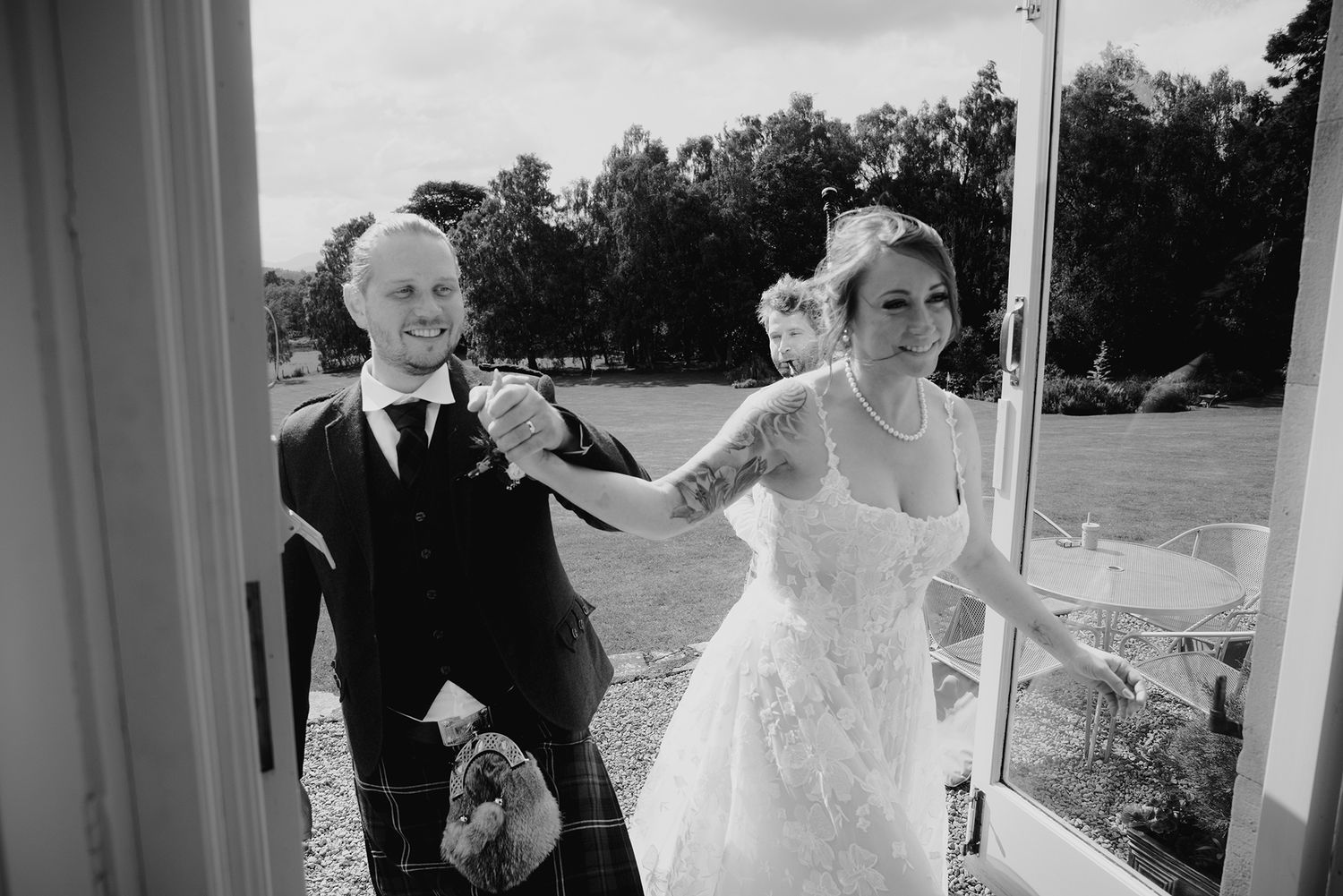 Black and white photo of smiling groom in kilt and bride in lace dress at wedding.