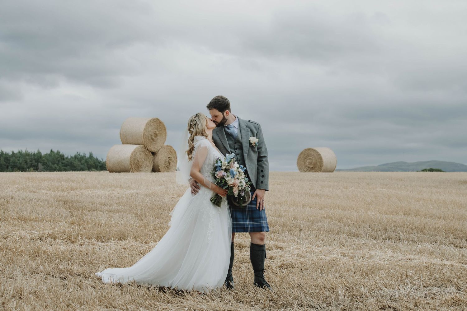 Bride and groom share romantic kiss in golden hay field with bales and mountains in background.