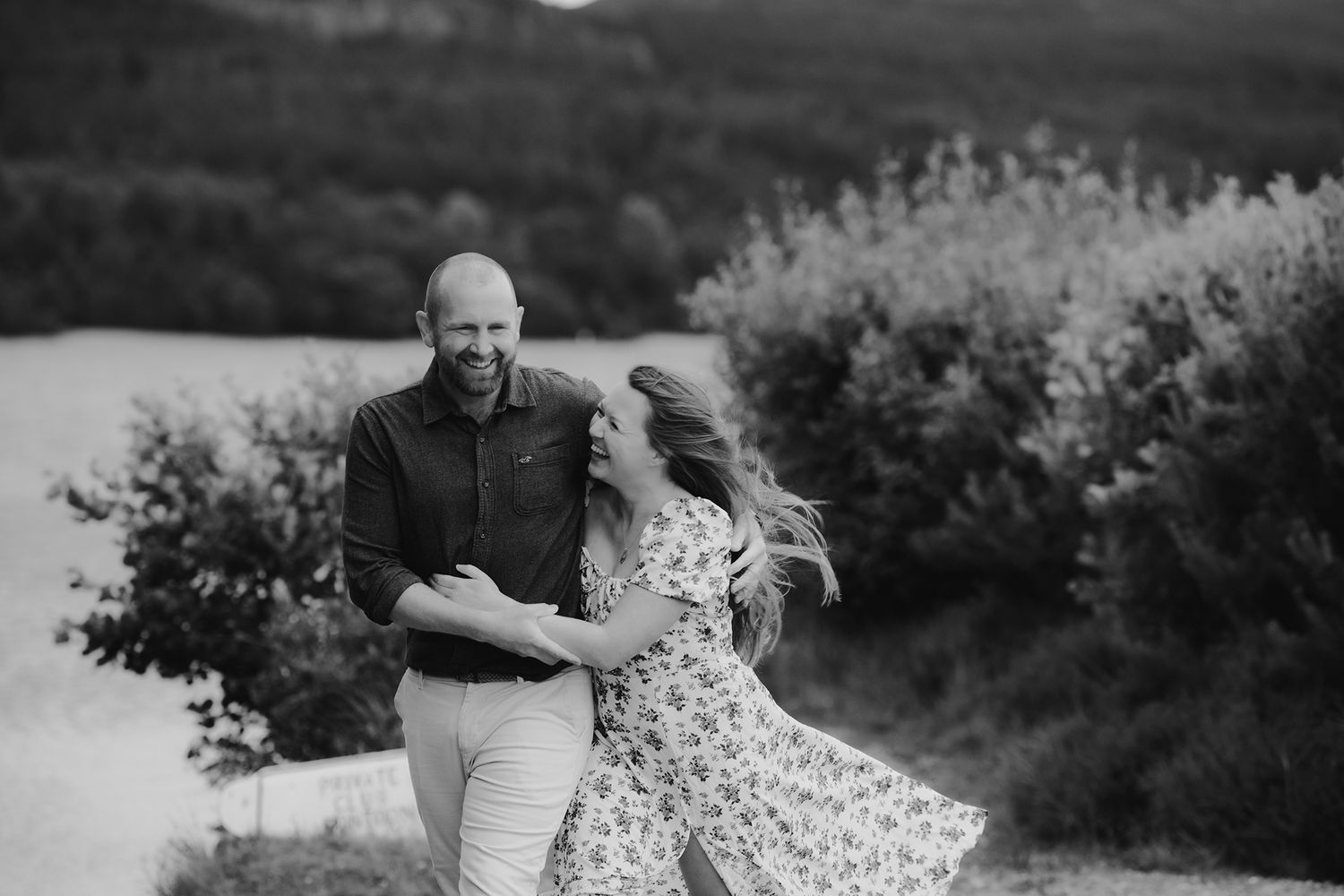 Black and white photo of a laughing couple embracing outdoors with wildflowers and trees behind them.