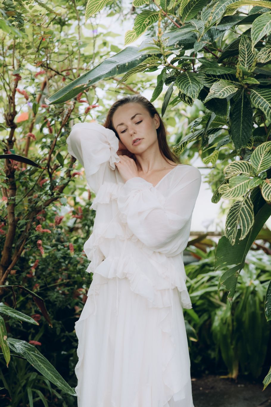 Woman in white ruffled long-sleeve dress with eyes closed among lush tropical greenhouse plants.