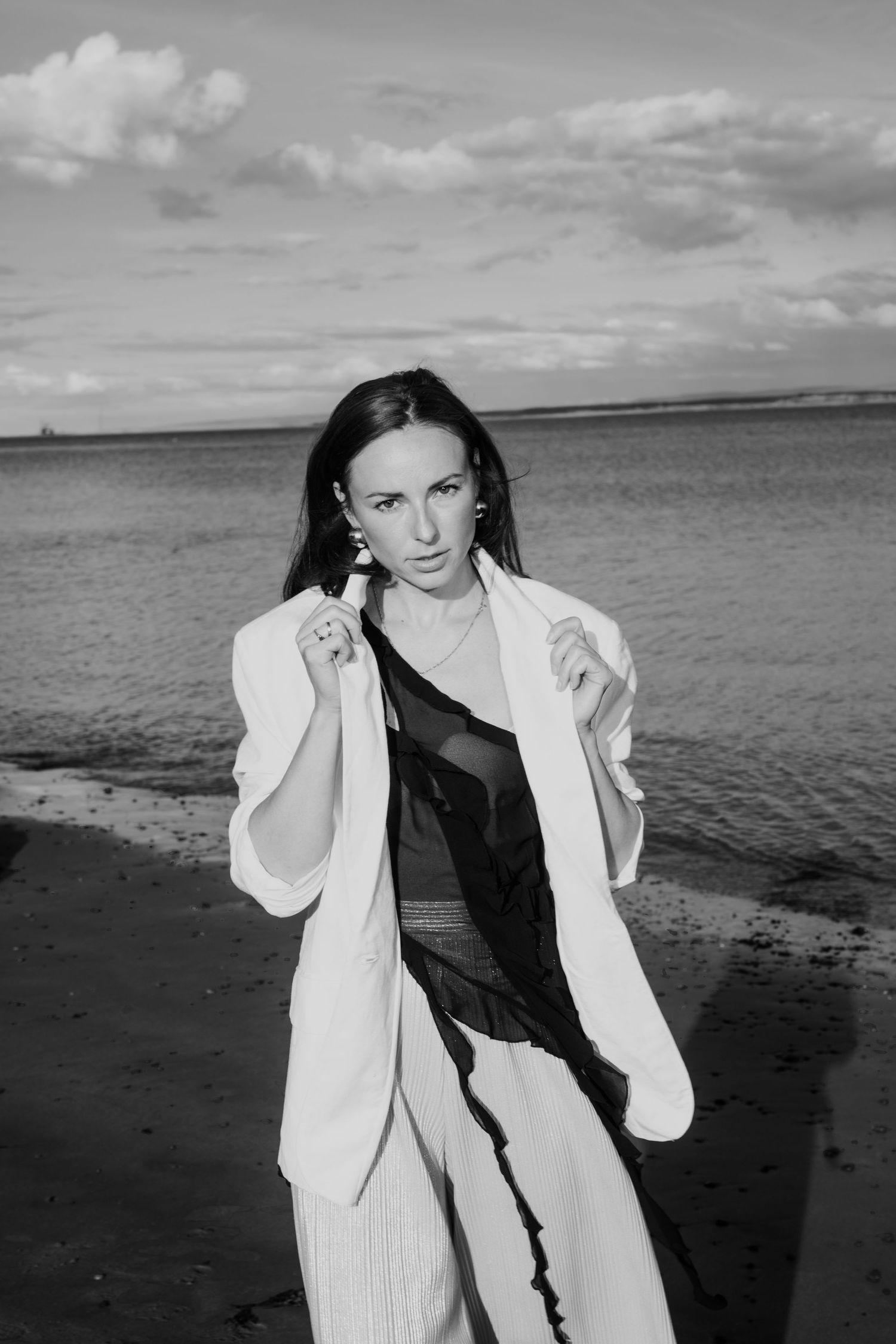 Black and white photo of a woman in white blazer standing on a sandy beach shoreline.