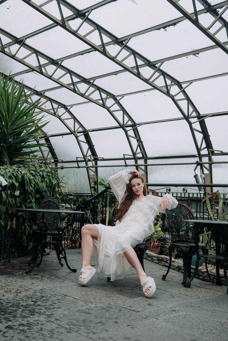 Woman in sheer white dress seated in ornate chair inside a glass-roofed Victorian greenhouse.
