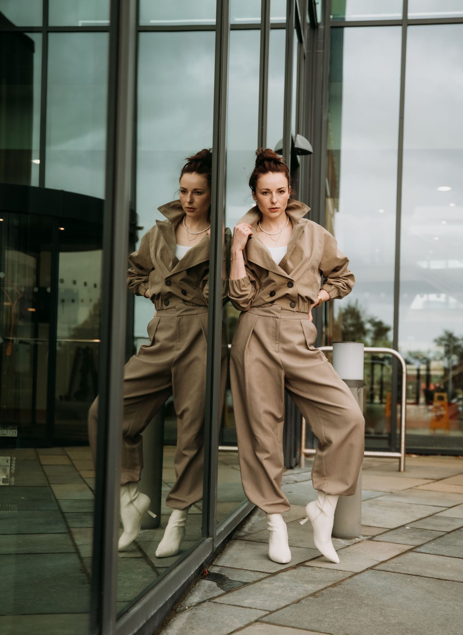 Woman in beige pantsuit posing against glass wall with mirror reflection outdoors.