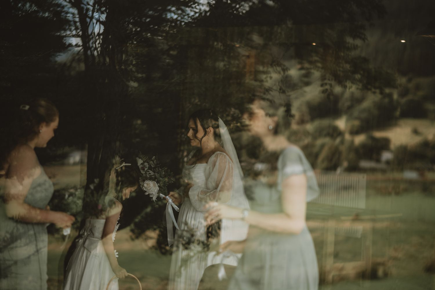 Artistic reflection of bride and bridesmaids in blue dresses captured through a glass window outdoors.
