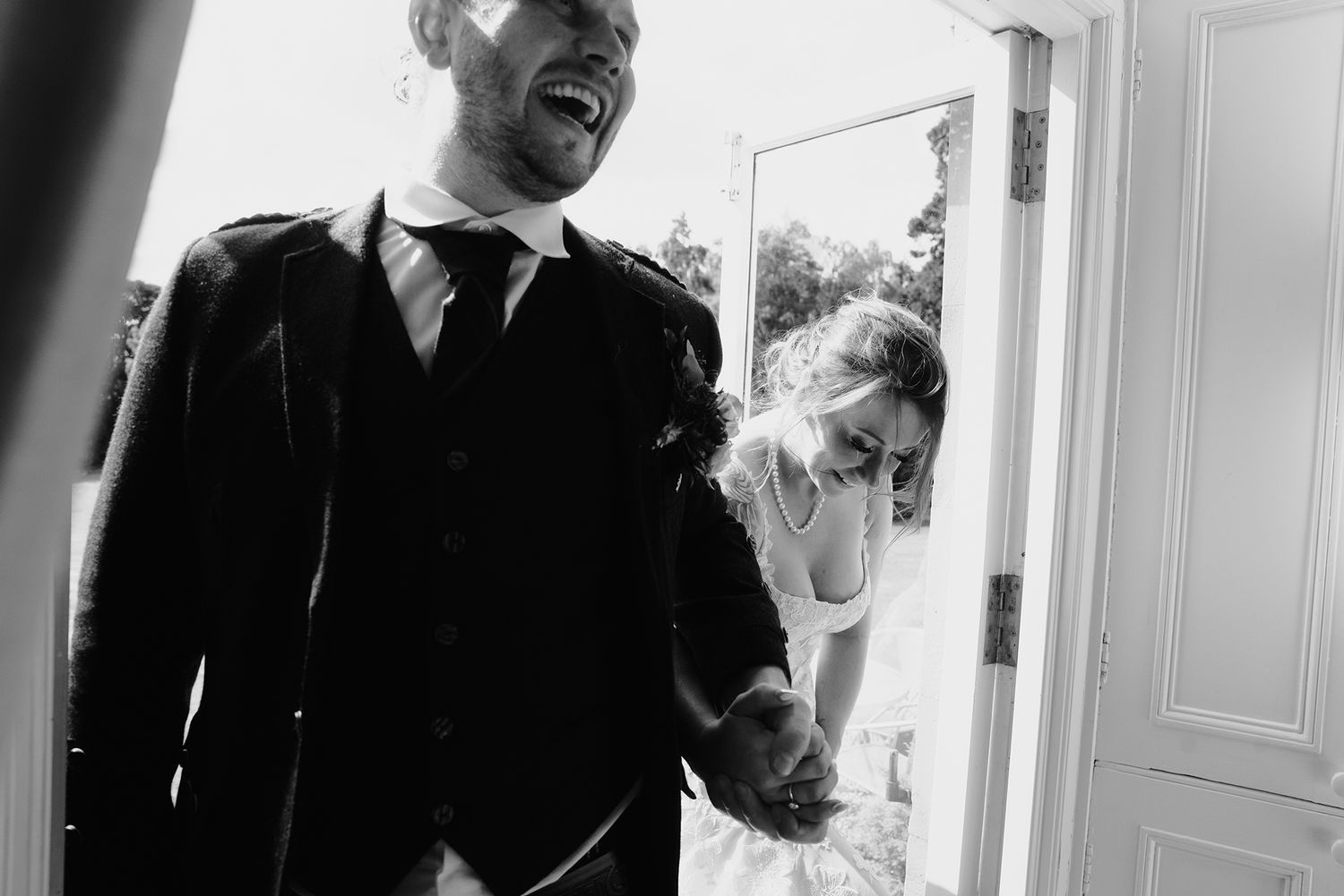 Black and white photo of laughing groom and bride holding hands at wedding venue door.