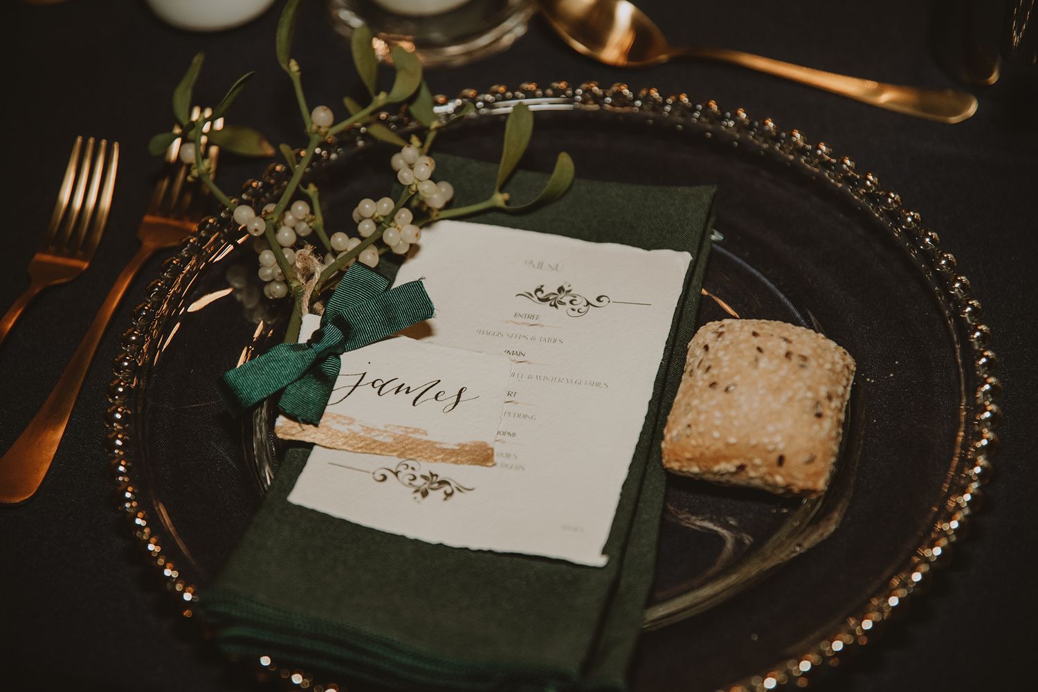 Elegant wedding place setting with green napkin, personalized menu card, gold cutlery, and floral sprig on charger plate.