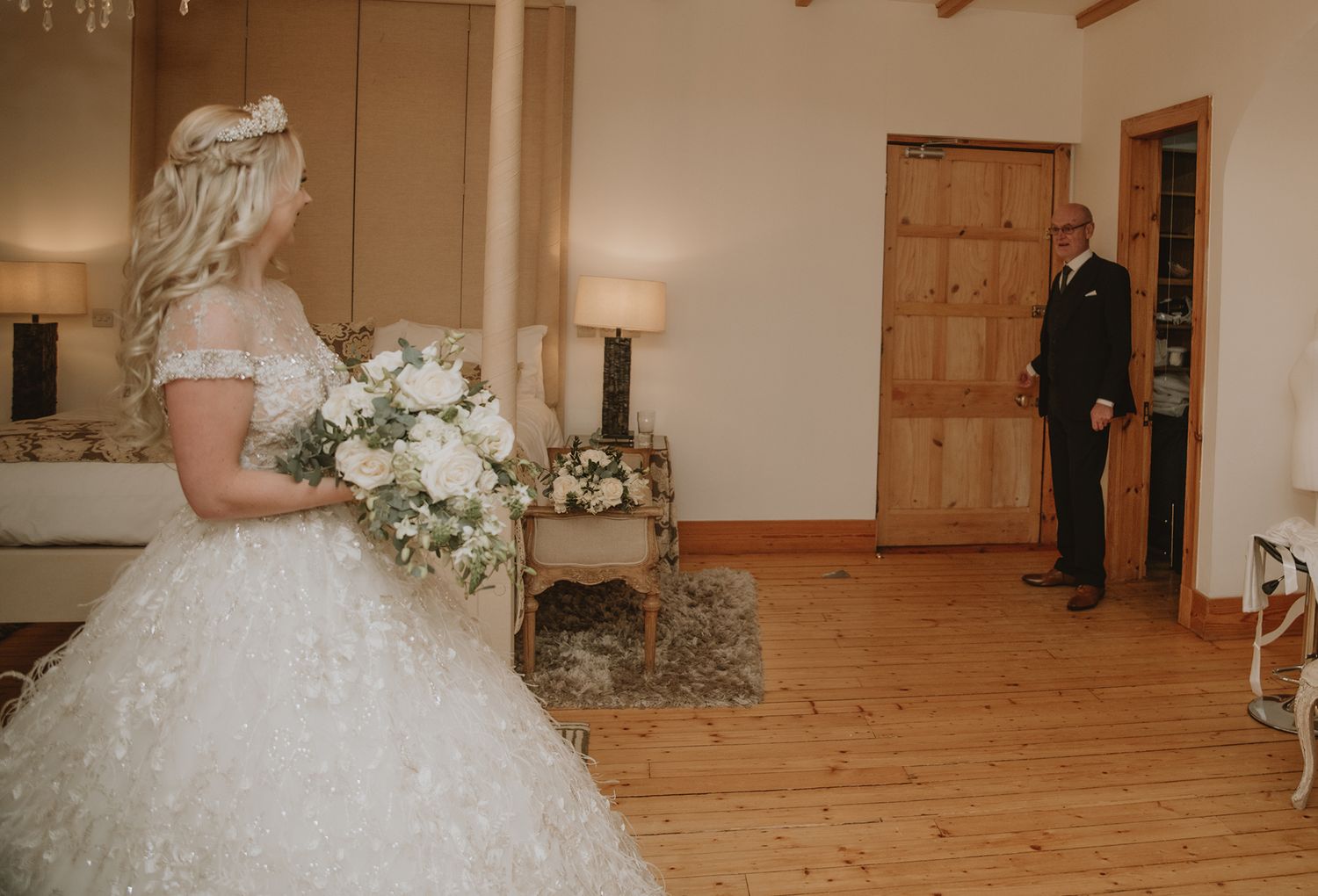 Bride in a white ball gown holding a floral bouquet stands in a rustic room as a man in a suit looks on.