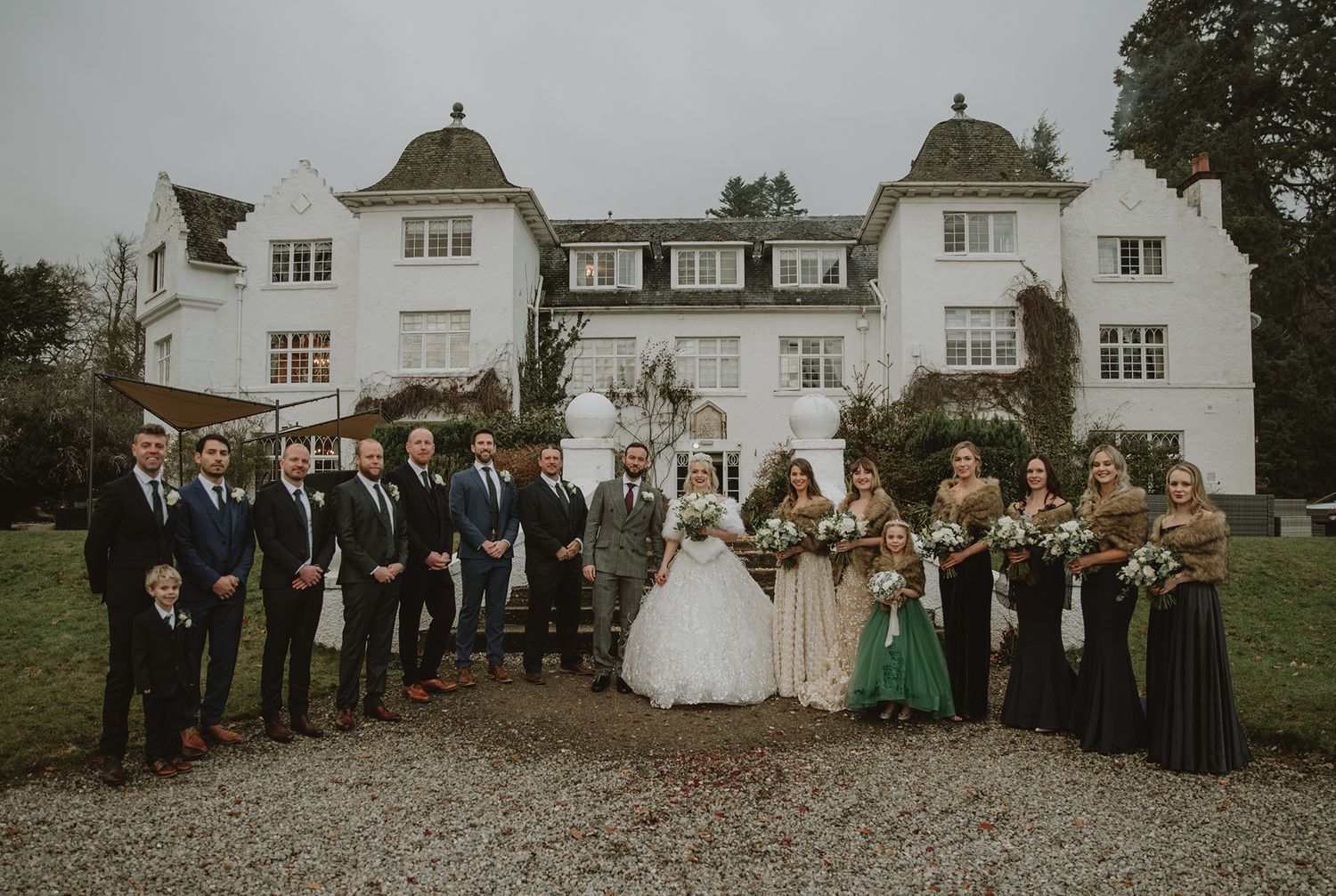 Wedding party poses in front of a white manor house, bride in white gown with groomsmen and bridesmaids in dark attire.