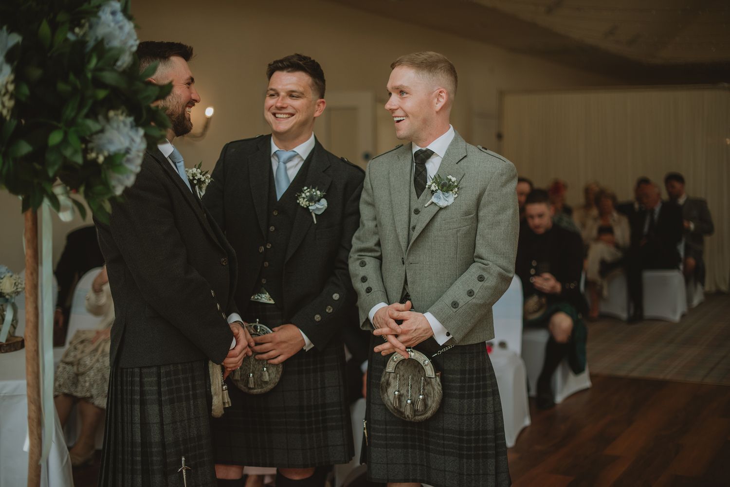 Three men in traditional Scottish kilts smiling during a wedding ceremony indoors.
