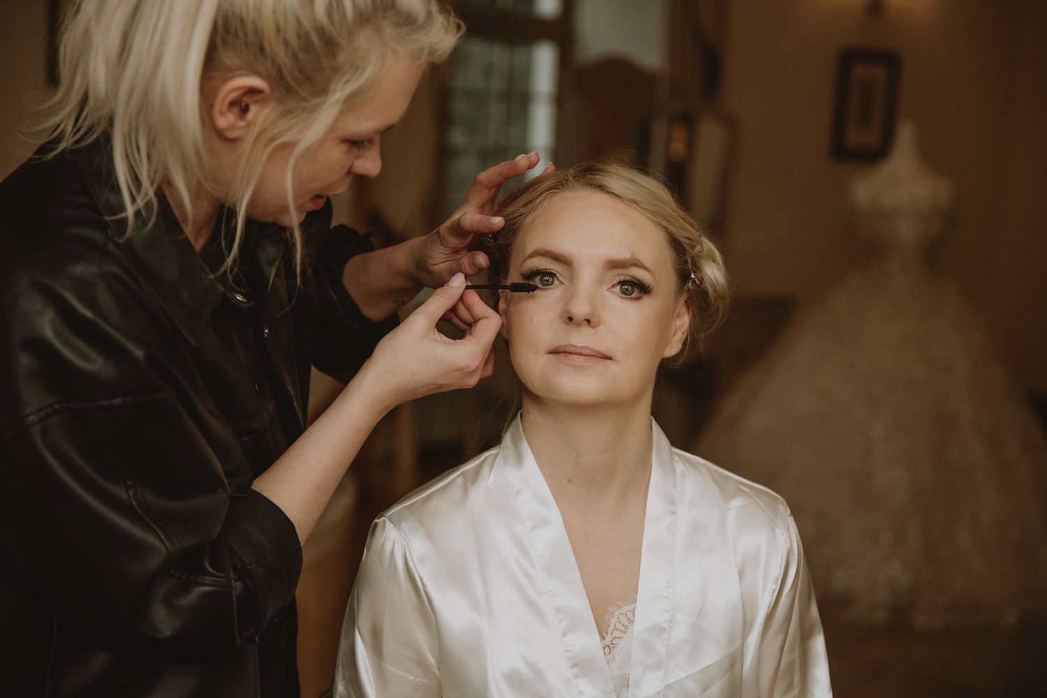Professional makeup artist applying eye makeup to bride wearing white satin robe on wedding morning.