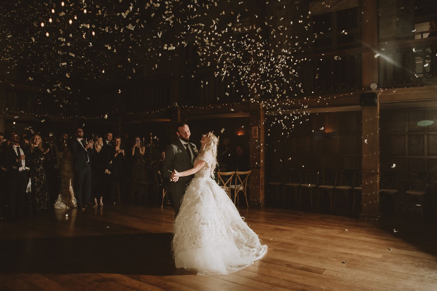 Bride and groom share first dance with confetti falling in a warmly lit rustic wedding venue.