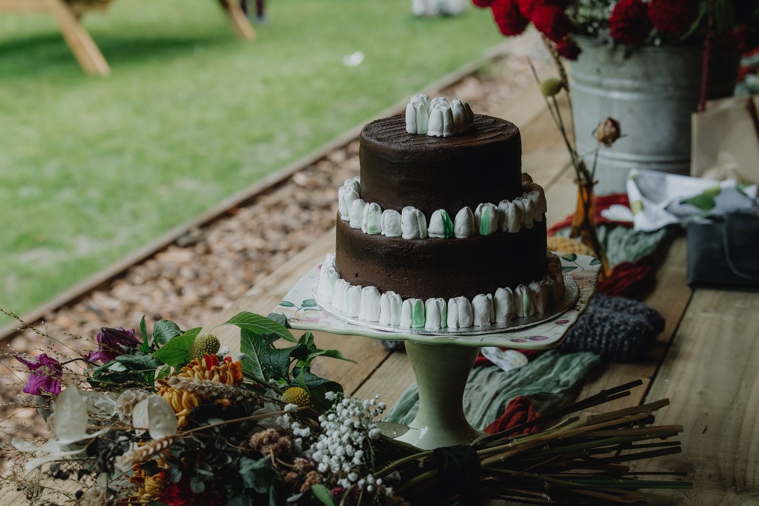 Two-tier chocolate wedding cake with macarons displayed on rustic wooden table with flowers.