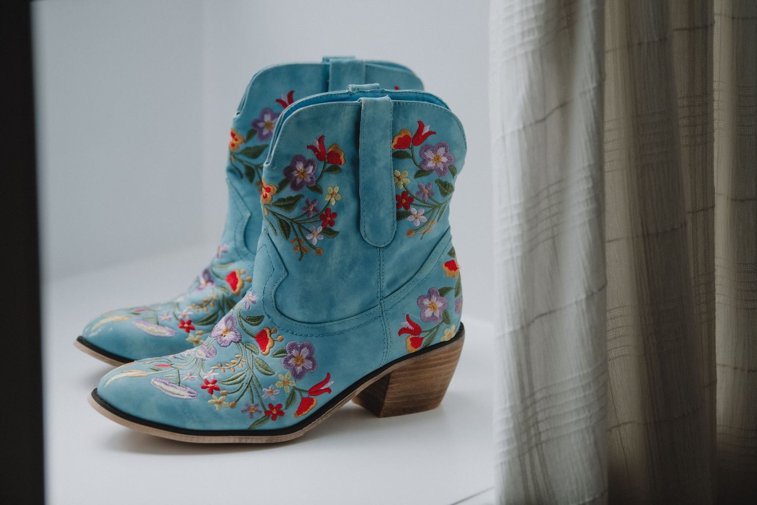 Turquoise cowboy boots with colorful floral embroidery displayed on a white windowsill.